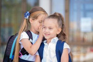 Two elementary-school-aged girls leaning in head-to-head as one whispers a secret in the other's ear, the kind of sustained hair contact that lets head lice crawl from one kid to another.