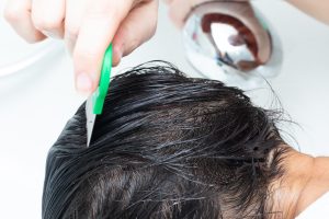 A parent's hands using a metal fine-tooth nit comb on a sectioned, shoulder-length child's hair under bright light, illustrating the manual comb-out step that has to happen after any lice treatment to remove eggs from the hair shaft.
