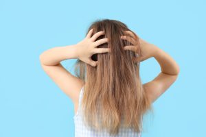Back view of a school-age child with medium-length hair reaching up to scratch the nape of the neck and the area behind the ears, illustrating the early scratching pattern parents often notice as a first sign of a head lice case.