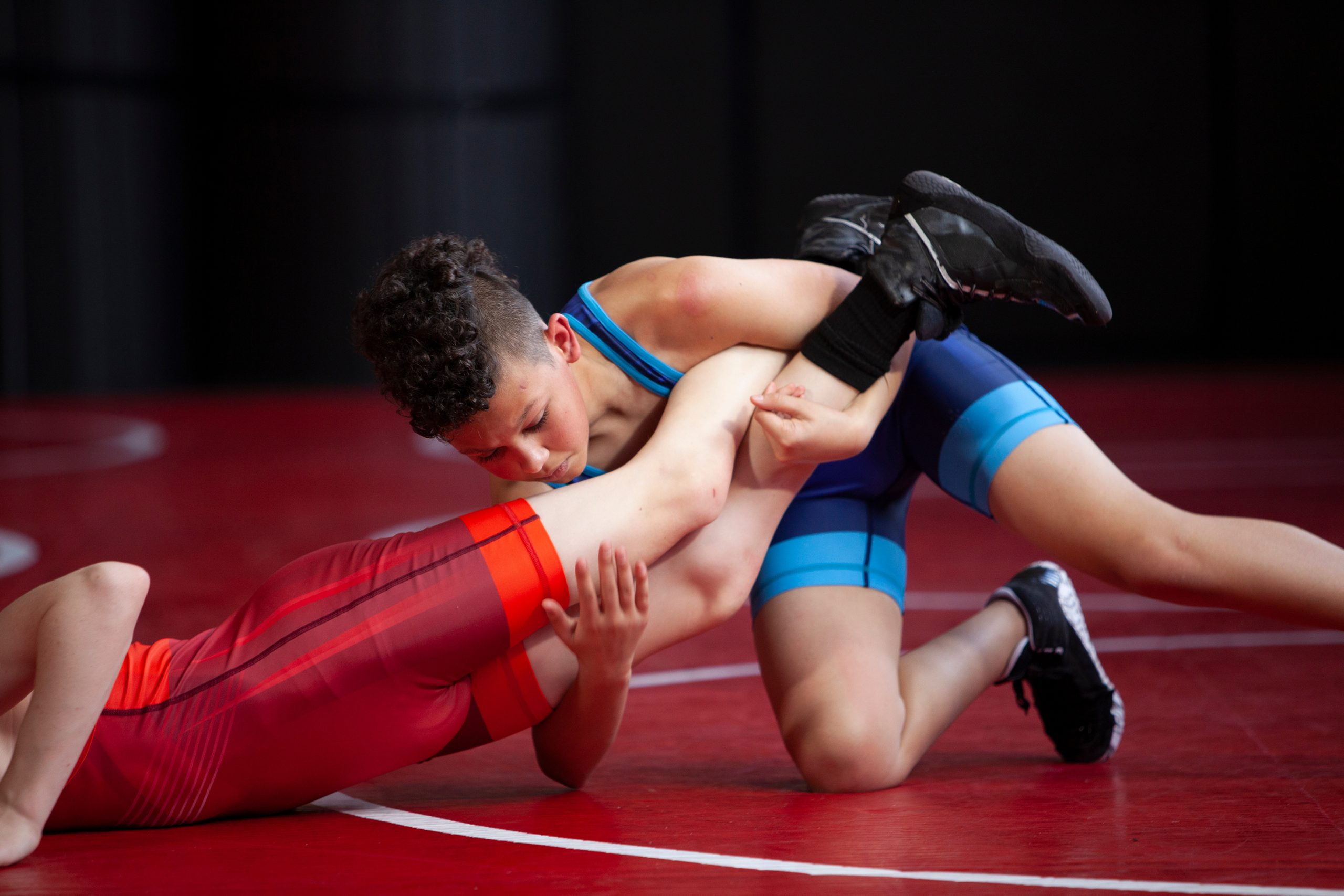 Wrestlers in red and blue singlets practicing on a red mat.