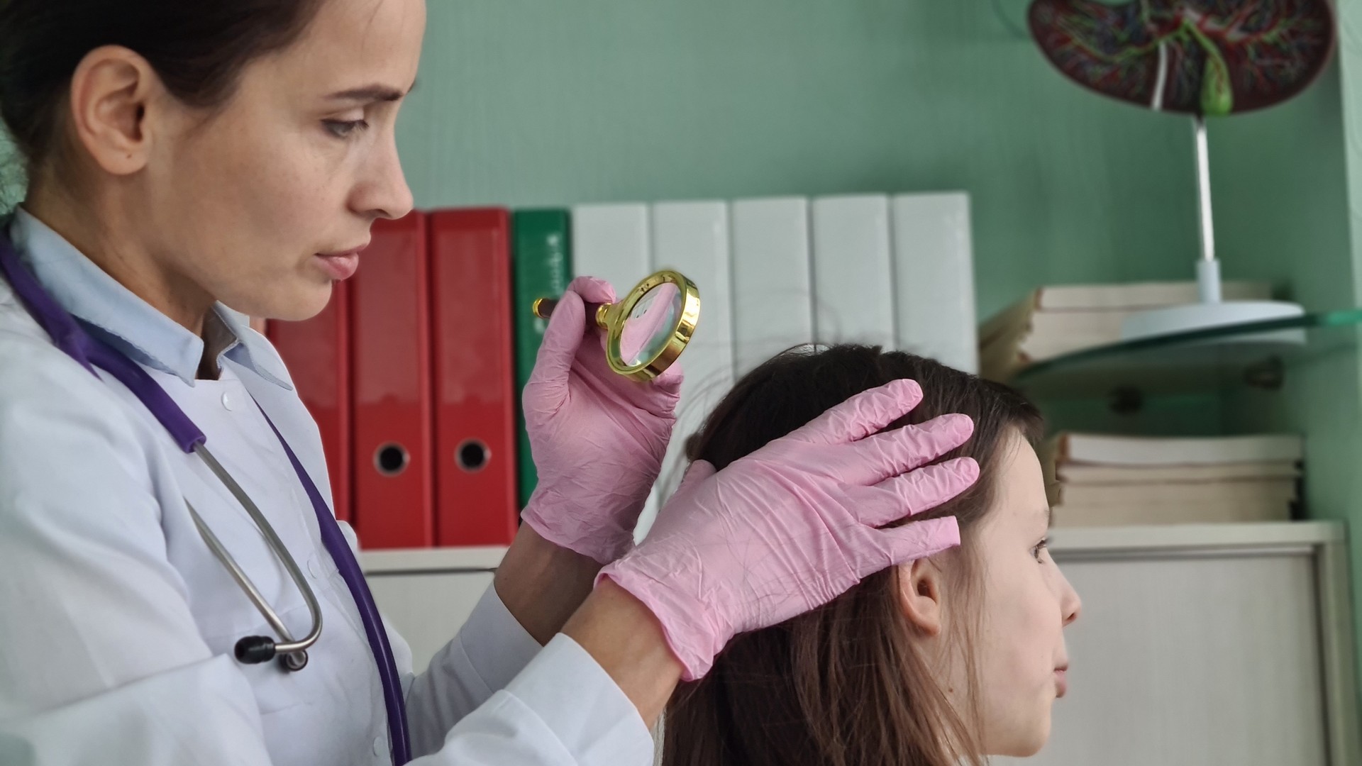 A lice-treatment technician parting a young child's hair into a section and examining the scalp under bright light with a fine metal comb during a professional head check at a clinic, the thorough check the article says home looks often cannot match.